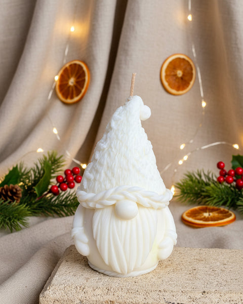 A white gnome figurine candle with a textured knit hat, braided beard trim, and long sculpted beard, displayed on a stone block against a burlap backdrop.