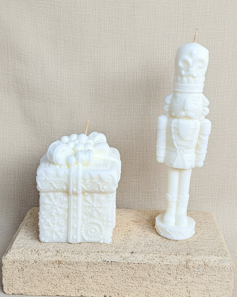 Two white novelty candles shaped like a gift box and a nutcracker soldier, displayed side-by-side on a textured concrete block against a burlap backdrop.
