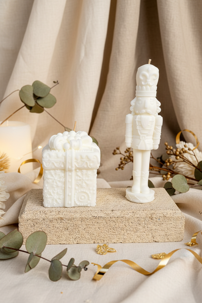 Two white novelty candles shaped like a gift box and a nutcracker soldier, displayed side-by-side on a textured concrete block against a burlap backdrop.