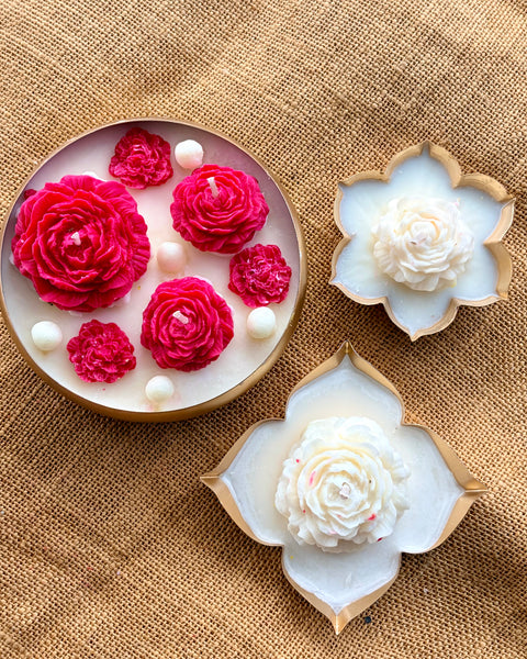 Three decorative flower-shaped candles arranged on a burlap surface: one round white candle with bright pink rose motifs and white pearl-like beads, and two white flower-shaped candles with gold-toned edges.
