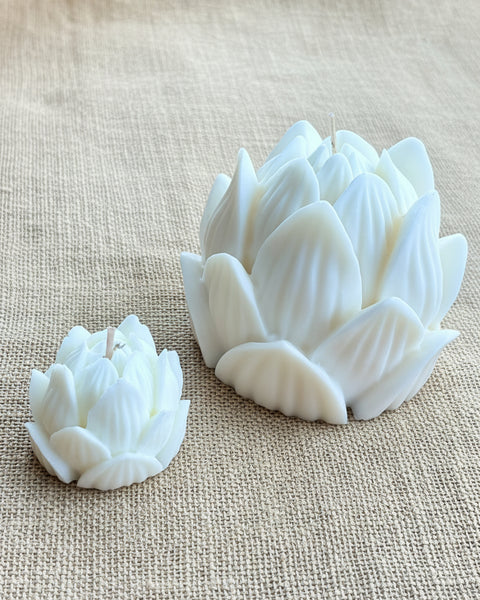 Two white lotus-shaped candles, one large and one small, resting on a textured beige burlap fabric background.