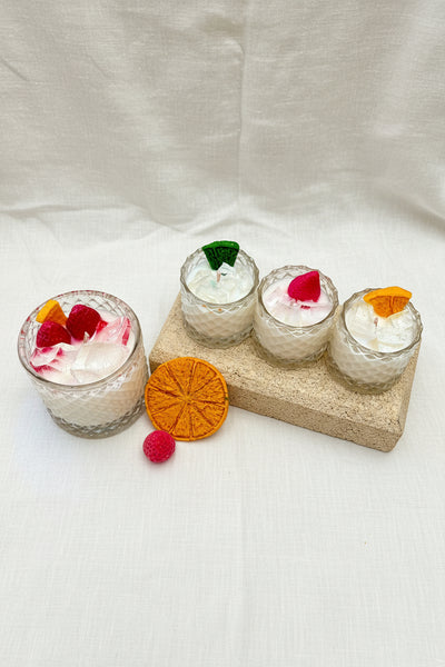 Four small glass jars with whipped cream, fruit slices, and a decorative stone block on a textured brown surface.