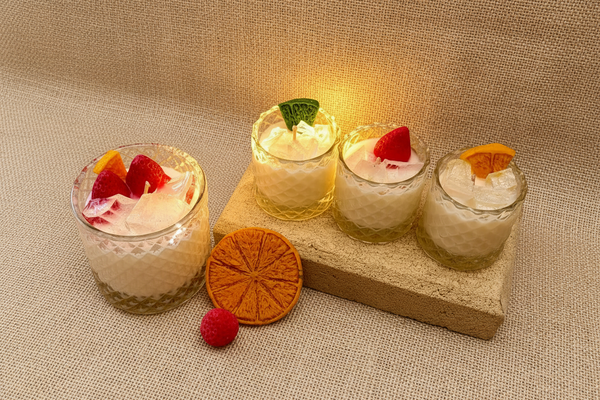 Four small glass jars with whipped cream, fruit slices, and a decorative stone block on a textured brown surface.