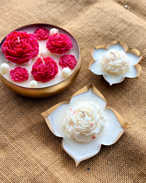 Decorative flower-shaped candles displayed on a burlap background, including a round gold dish with bright pink rose candles and two white lotus-shaped candles with gold edges.