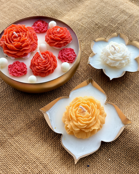 Decorative flower-shaped candles displayed on a burlap background, including a round gold dish with bright pink rose candles and two white lotus-shaped candles with gold edges.