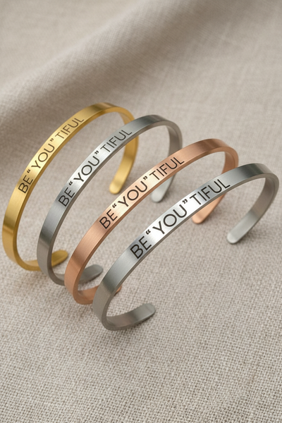 Four 'BE YOU TIFUL' bracelets in gold, silver, rose gold, and bronze on a textured surface.