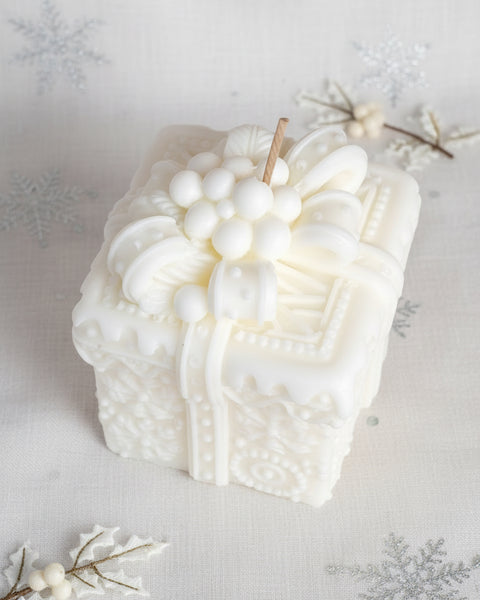A white decorative candle shaped like a gift box, featuring embossed floral and berry details on top, photographed on a textured burlap background.