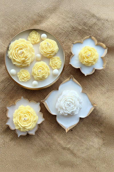 Four flower-shaped candles arranged on a textured beige fabric background. One round candle holder contains multiple pale yellow rose-shaped candles with white pearl-like accents, while three other holders contain single white and pale yellow rose candles with gold-toned edges.