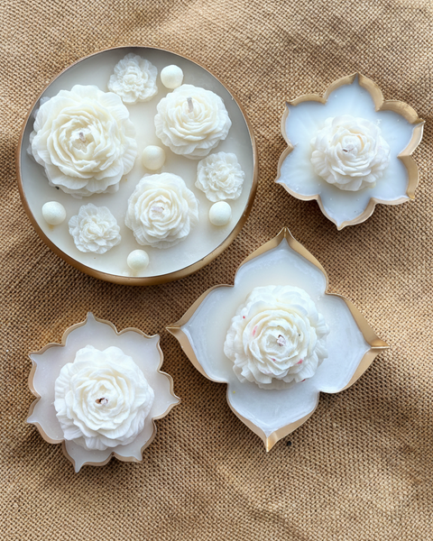 Four decorative flower-shaped candles arranged on a beige burlap background. One round candle is bright pink with five large rose blooms and small white pearl-like accents.