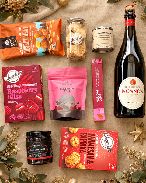 A curated gift assortment of snacks and beverages arranged on a burlap fabric background, including a bottle of Nonno's Prosecco, bags and jars from Charlie's and Sesh, and boxes from Goodies.