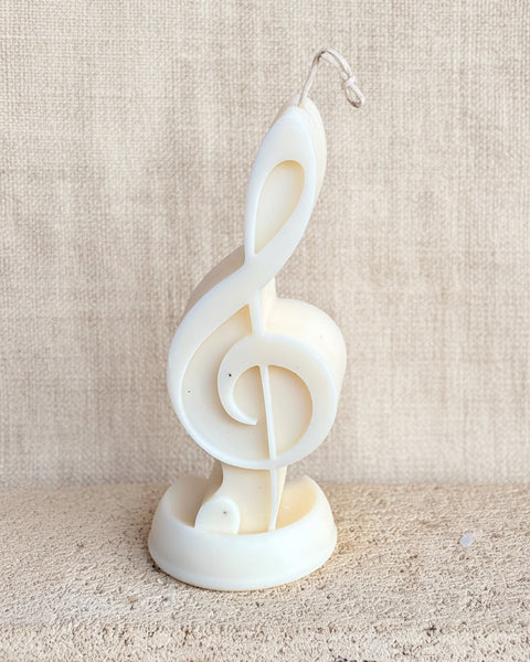A white novelty candle sculpted in the shape of a treble clef, resting on a textured stone block against a rustic burlap backdrop.