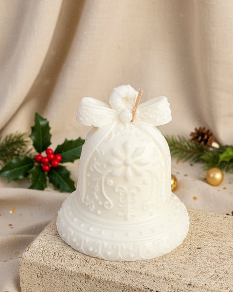 A white decorative bell-shaped candle with embossed floral and scroll patterns, topped with a bow, displayed on a stone block against a burlap backdrop.