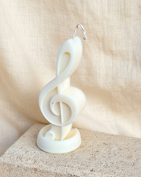 An off-white novelty candle shaped like a treble clef, standing on a textured stone block against a rustic burlap backdrop.
