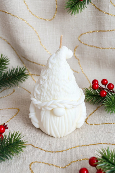 A white gnome-shaped candle with a textured hat, braided beard detail, and a single wick, photographed on a tan burlap fabric background.