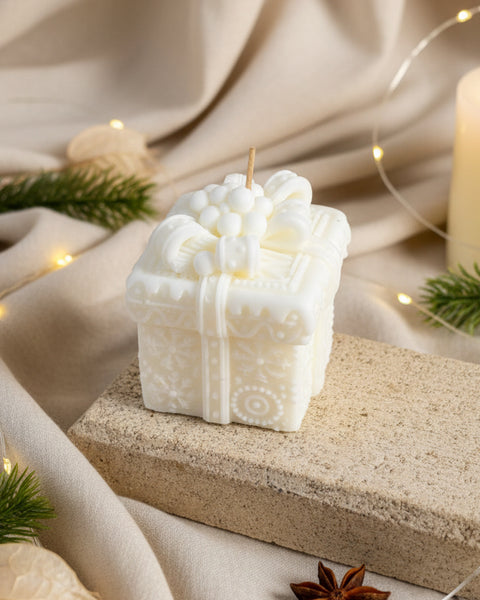 A small white decorative candle shaped like a gift box with a floral and berry design on top, displayed on a stone slab against a burlap background.
