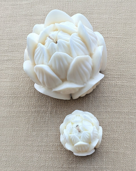 Two white lotus-shaped candles, one large and one small, displayed on a textured tan burlap fabric background.