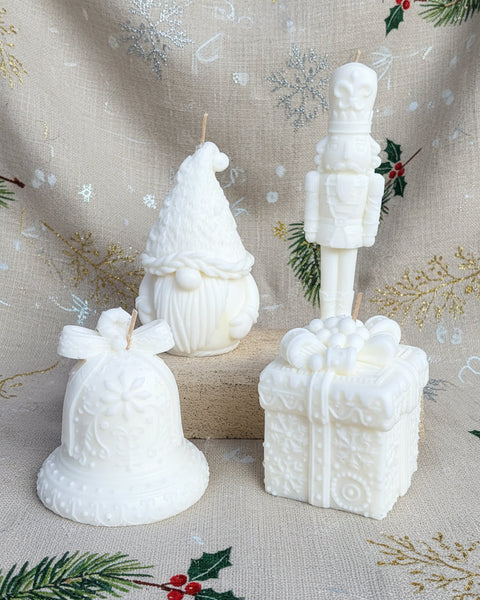 Three white holiday-themed sculptural candles on a burlap background: a gnome candle, a nutcracker candle, and a gift box candle.