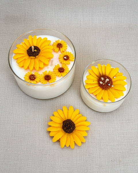 Two small glass containers with yellow flower-shaped candles on a textured brown fabric background.