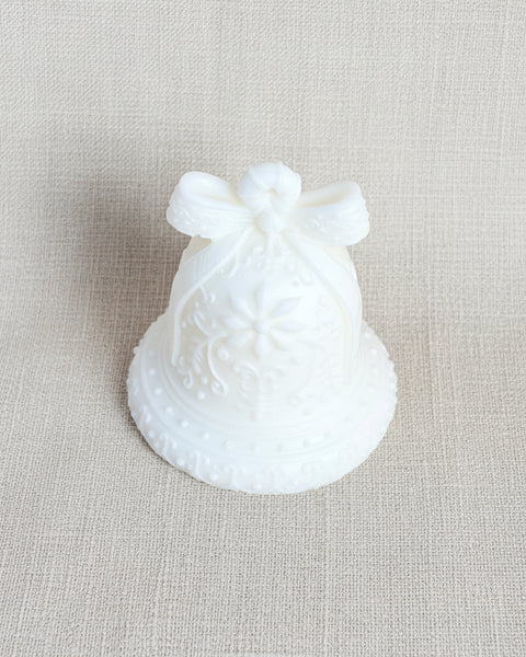 A small white decorative bell-shaped candle with a bow on top, placed on a textured tan burlap fabric background.