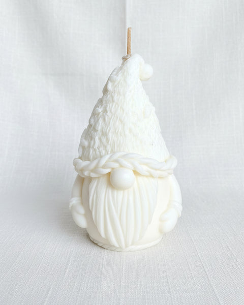A white gnome-shaped candle with a textured hat, braided scarf detail, and long beard, photographed against a burlap fabric background.