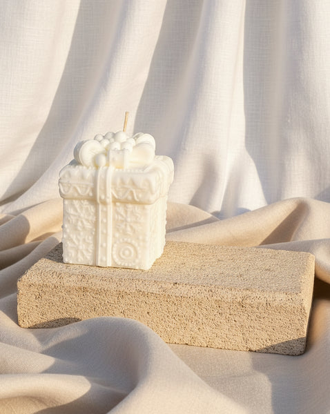 A white gift box candle with embossed floral patterns and a wick on top, displayed on a rectangular stone block against a burlap backdrop.