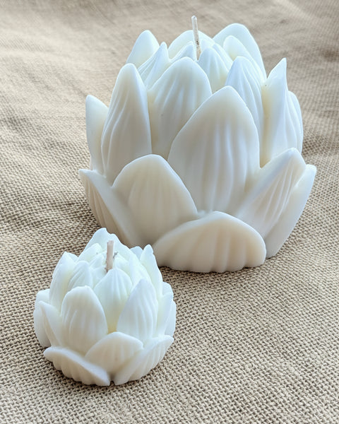 Two white lotus-shaped candles, one large and one small, displayed on a textured tan burlap fabric background.