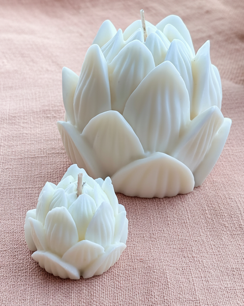Two white lotus-shaped candles, one large and one small, displayed on a textured dusty pink fabric background.