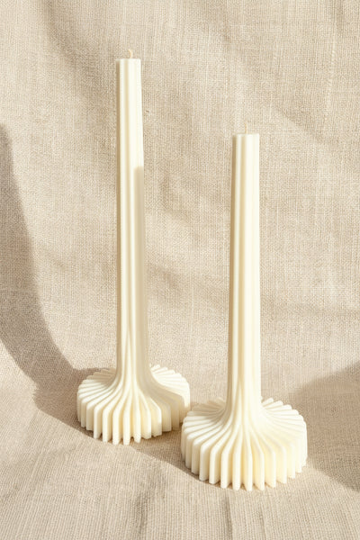 Two tall white candle holders with fluted circular bases, standing on a textured tan burlap background.