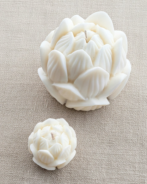 Two white lotus-shaped candles, one large and one small, displayed on a textured tan burlap background.