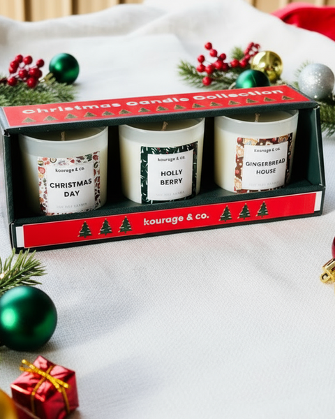 Set of three scented candles in a decorative box with Christmas-themed labels, surrounded by festive decorations.