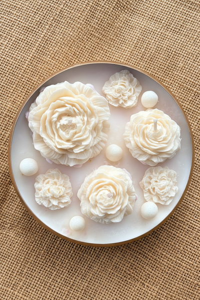 A round white candle with a gold-toned rim, decorated with five vibrant pink flower candles and five small white beads, set on a textured burlap background.