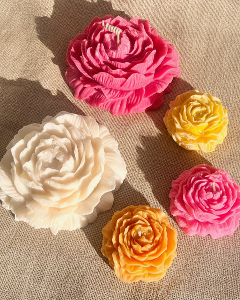 Four flower-shaped candles in pink, white, yellow, and orange on a textured fabric background.